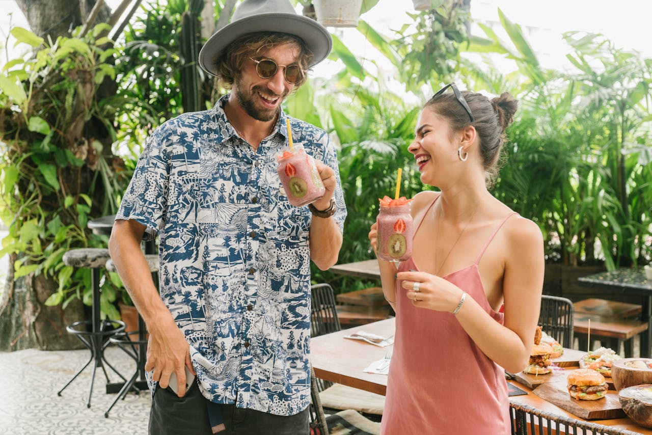 A joyful couple laughs together while enjoying tropical fruit smoothies at an outdoor cafe surrounded by lush greenery.