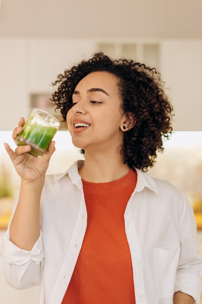 Smiling woman with curly hair enjoying a fresh green smoothie indoors, highlighting a healthy lifestyle.
